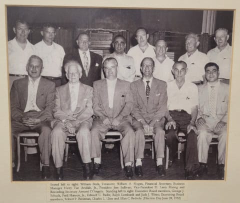 An old black-and-white photo of 15 men, including executives and members, gathered for a formal grou.