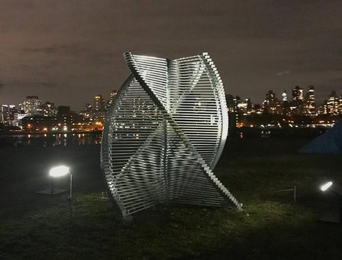 Sculpture of intertwined metal slats illuminated against city skyline backdrop.